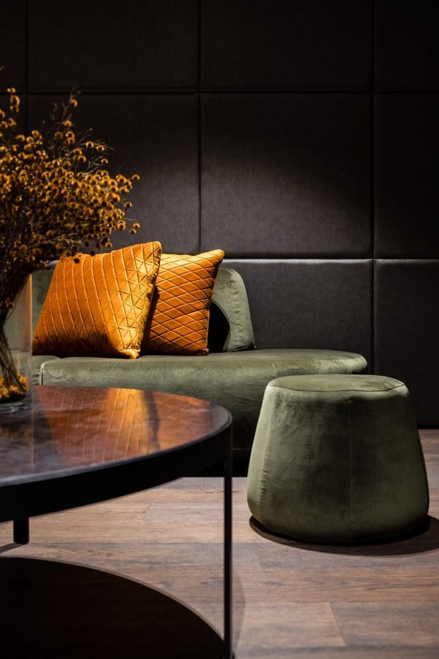 A moody, luxurious living room vignette featuring a low green velvet sofa and matching rounded ottoman, orange quilted cushions, a dark padded wall, a dark wood and glass coffee table, and warm timber flooring. Dried botanical branches add a small decorative accent.