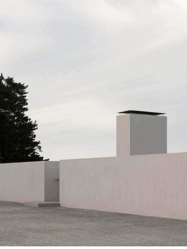 Minimalist exterior view of a low, pale rendered compound wall with a tall rectangular tower rising behind it. The tower has a thin projecting flat roof with a dark underside, while a recessed pedestrian entry is visible in the wall. A gravel forecourt and mature evergreen tree sit against an overcast sky, creating a restrained, monolithic composition.
