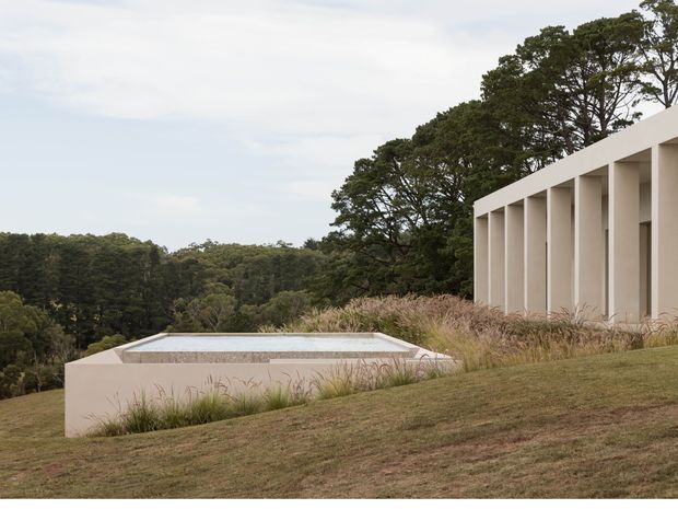 Exterior architectural view of a minimalist, low-profile residence set into a gently sloping landscaped site. A long pale rendered structure with a repetitive colonnade or deep vertical openings stands to the right, while a shallow rectangular reflecting pool or water feature is integrated into the foreground terrace. Ornamental grasses and mature woodland surround the building, creating a restrained, natural setting.
