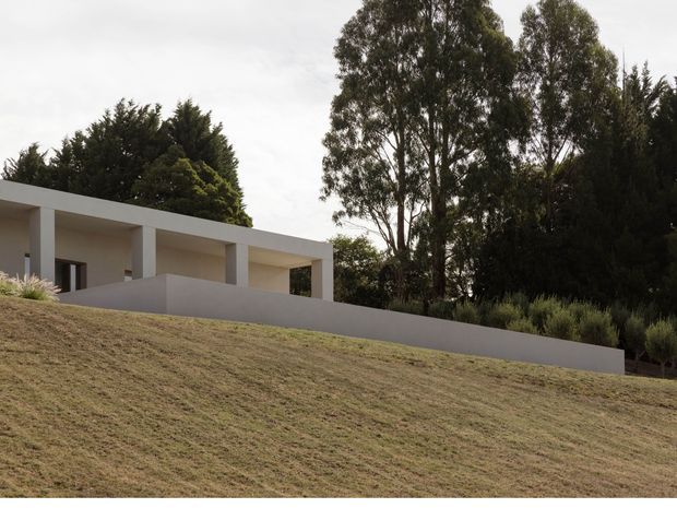 A low, minimalist white house set into a gently sloping, landscaped site. The building has a long flat roof, deep covered outdoor areas, square structural columns, broad parapet walls, and restrained rectangular openings. Mature trees and shrubs form a dense backdrop, while the foreground is a closely mown grassy slope.