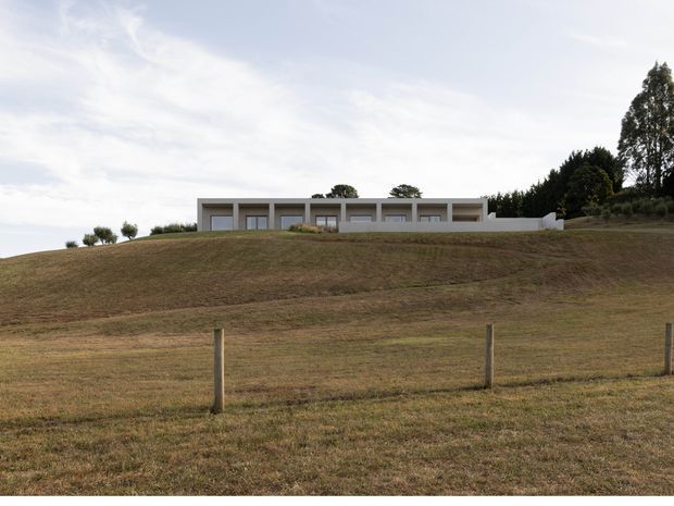 A long, low modern residence viewed from across a gently sloping rural field. The building has a minimalist white facade with a strong horizontal roofline, regularly spaced recessed openings, and extensive low planting and trees around it. A simple wire-and-post fence crosses the foreground beneath an expansive pale sky.