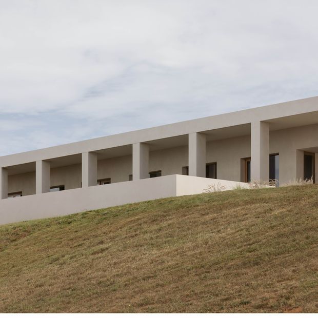 A minimalist, elongated low-rise building viewed from below across a sloping grass-covered site. The pale rendered facade features a strong horizontal roofline, a repeated rhythm of deep rectangular openings supported by slender square columns, and dark-framed windows and doors set within recessed bays. The overcast sky and sparse landscaping create a restrained, contemporary architectural composition.