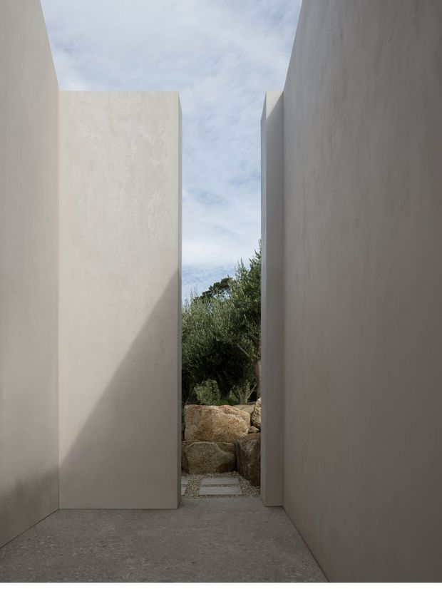 A narrow architectural passage framed by tall, smooth pale rendered walls, opening toward a landscaped rocky garden with olive-like planting. The space has a minimalist monolithic character, with a grey textured paved or concrete floor, strong geometric planes, and a vertical slice of sky above.