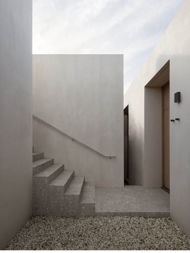 Minimalist architectural courtyard passage with pale rendered walls, a sculptural terrazzo or concrete stair, gravel ground, and a simple timber-toned recessed doorway. The composition is geometric and softly lit, with an open sky view above and restrained black wall fixtures.