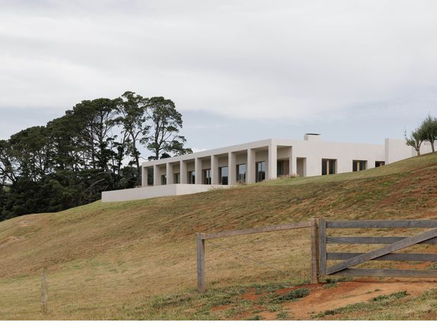 A low, minimalist modern house set into a gently sloping grassy landscape. The pale rendered building has a long rectilinear form, deep recessed openings and a repeated colonnade along the front, with a flat roof and sparse surrounding planting. A timber farm-style gate and fence occupy the foreground, while mature trees sit behind the house under an overcast sky.