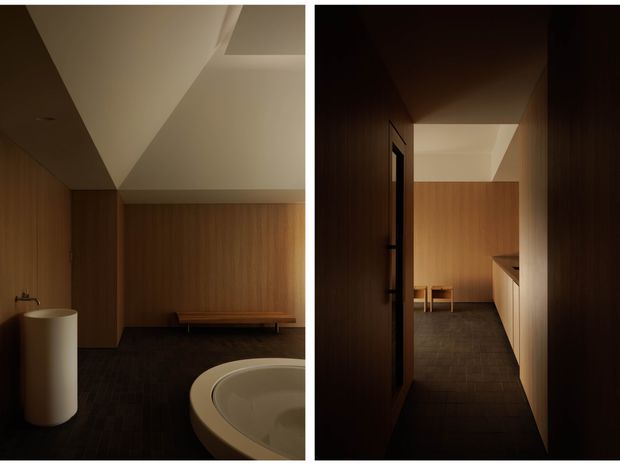 Diptych interior view of a dark, refined bathroom and adjoining circulation/vanity area. Warm timber wall panelling and cabinetry contrast with dark tiled flooring, a freestanding cylindrical basin, and a partially visible rounded freestanding bathtub. The right panel shows a narrow hallway or entry into the bathroom with built-in cabinetry and a softly illuminated angular ceiling.