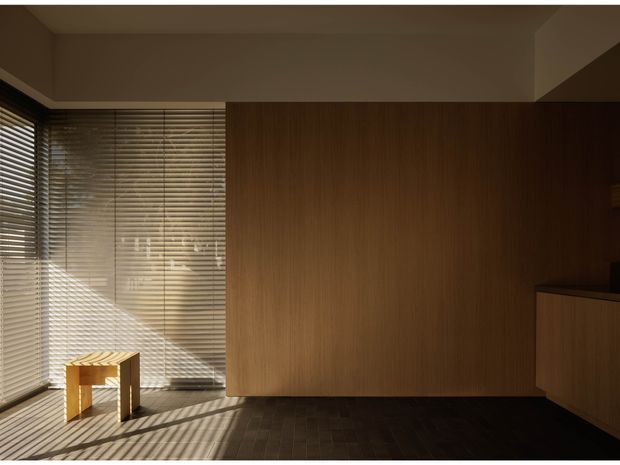 A warm, minimalist interior corner with a floor-to-ceiling corner window fitted with horizontal blinds, a dark timber-lined wall and matching built-in cabinetry, and dark timber flooring. Strong afternoon sunlight creates striped shadows across the floor, with a small light timber stool positioned by the glazing.