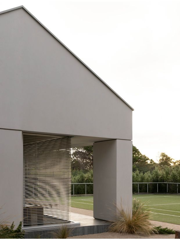 Exterior architectural view of a minimalist contemporary structure with a simple gabled roof form, smooth light-grey rendered walls, and a large covered opening supported by a square column. Horizontal metal screening creates a semi-transparent enclosure at the lower level. The building overlooks a fenced green sports court, with ornamental grasses and trees surrounding the site.