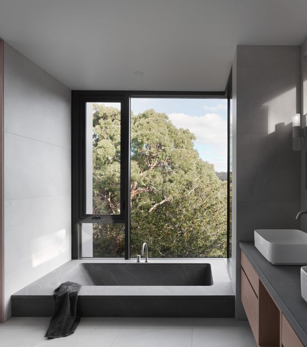 Contemporary bathroom with a deep dark freestanding-style built-in bathtub positioned beneath a large black-framed corner window overlooking trees. The room has pale large-format wall and floor tiles, a dark stone-look vanity benchtop, timber-toned vanity cabinetry, white vessel basins, and minimalist chrome tapware. Natural light creates a calm, refined atmosphere.