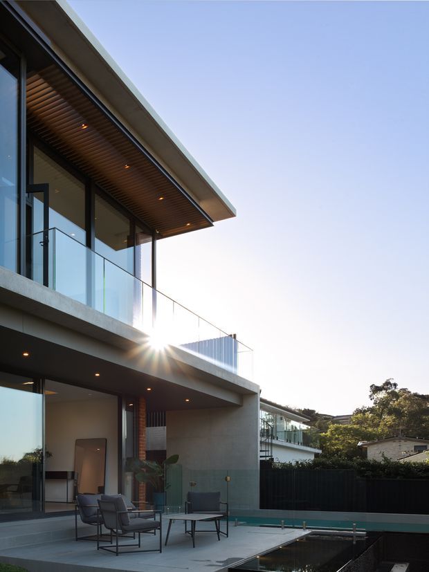 Photograph of a contemporary multi-level residence viewed from an outdoor terrace. The house features broad concrete cantilevered slabs, extensive floor-to-ceiling glazing with dark slender frames, a clear glass balustrade, and a projecting roof with a warm timber-lined soffit and recessed downlights. Outdoor lounge furniture sits on a paved terrace beside a narrow reflecting pool or swimming pool, with landscaped suburban surroundings visible beyond.