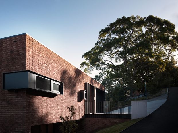 Exterior view of a contemporary brick building with a long, low rectilinear form. The facade features projecting dark-framed windows, a recessed entry or opening with vertical timber-like screening, and a raised walkway with metal railings. A sloping driveway and mature tree occupy the surrounding site.