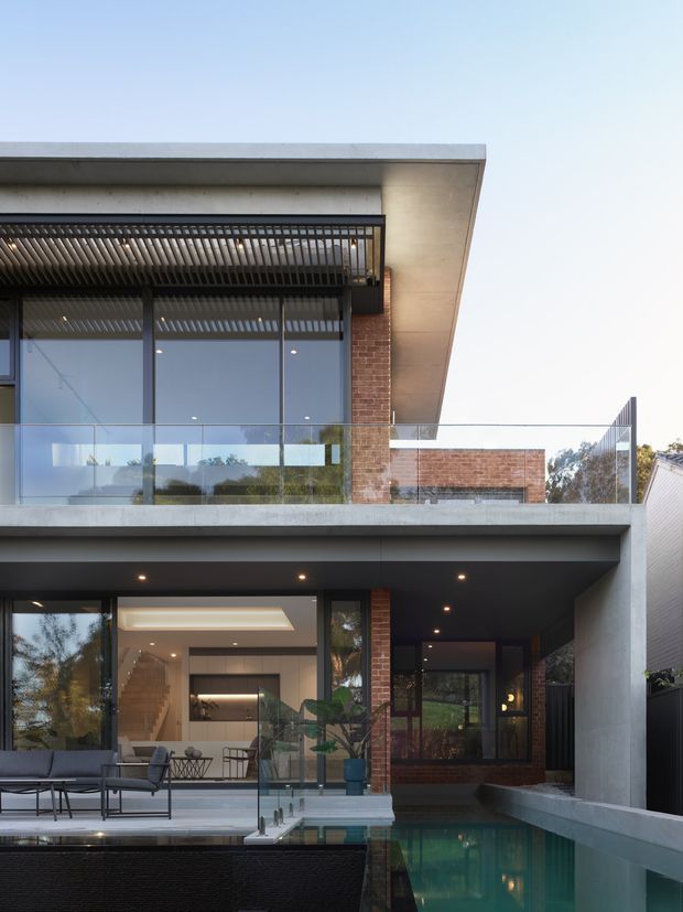 Contemporary multi-level residential facade with extensive floor-to-ceiling glazing, exposed red brickwork, concrete slab edges, a deep covered terrace, and a narrow reflecting or swimming pool alongside the ground floor. A rooftop-style pergola with vertical timber or metal battens shades the upper balcony, while glass balustrades and integrated recessed lighting reinforce the modern architectural character.