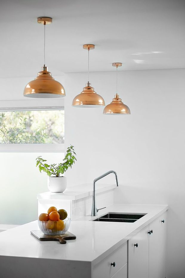 Bright compact white kitchen with a peninsula or island bench, integrated black double-bowl sink, curved chrome mixer tap, white fixed cabinetry, and a white tiled backsplash. Three copper-toned pendant lights hang above the work surface, while a large window brings in natural light. A potted plant and fruit bowl add small decorative touches.