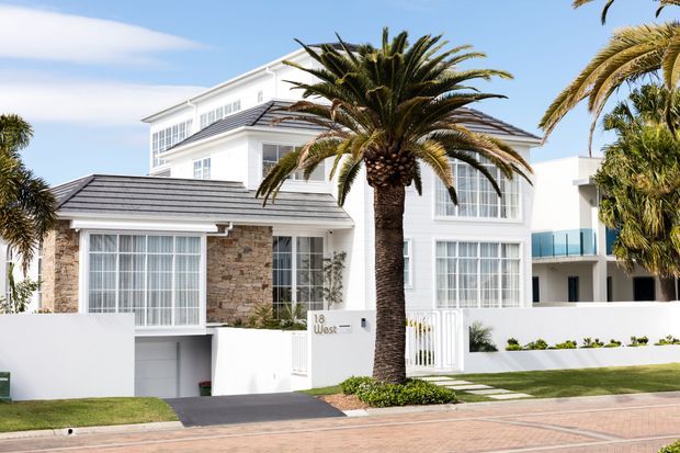 Bright exterior view of a large white multi-storey coastal-style residence with dark tiled pitched roofs, extensive white-framed glazing, a stone-clad projecting wing, balconies, white boundary walls, manicured lawn and mature palm trees. The property is viewed from the street and has a clean, resort-like presentation.