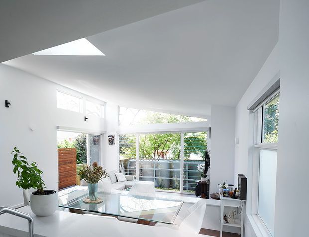 Bright, contemporary open-plan living and dining area with white walls and ceiling, extensive glazing, a skylight, and views to leafy vegetation outside. A glass dining table with white chairs occupies the foreground, while a small seating area and decorative plants sit near the windows. The space has a clean, airy architectural character with strong natural light and angular ceiling geometry.