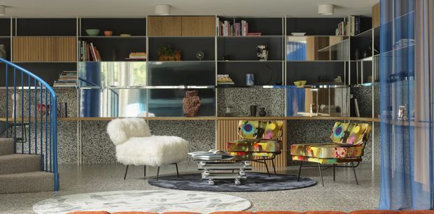 Eclectic contemporary living room with a dramatic full-width built-in shelving and display wall, terrazzo-clad lower cabinetry, a curved blue stair railing, sculptural boucle armchair, colourful patterned lounge chairs, layered rugs, and a low stacked coffee table. Floor-to-ceiling blue curtains frame the right side, while reflective surfaces and bright accents add a playful, gallery-like character.