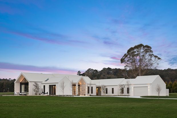 A wide exterior view of a low, sprawling white residential building set in a large lawn. The home is composed of several connected gabled volumes with crisp white cladding, dark-framed windows, covered outdoor areas, and an integrated double garage. A wooded backdrop and colourful dusk sky frame the property.