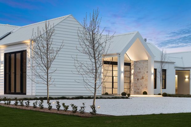 Exterior view of a large contemporary white-clad residence at dusk, featuring multiple pitched gabled roof forms, vertical black-framed glazing, a prominent illuminated entry canopy, and a pale natural-stone feature wall. The building is set within a carefully landscaped foreground with white gravel, clipped shrubs, young trees, and lawn.