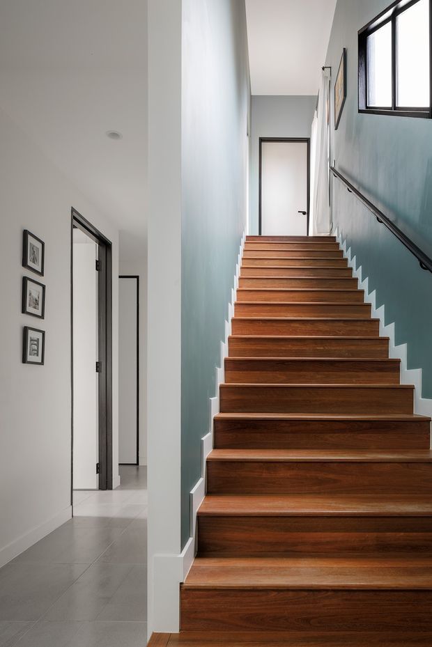 A bright, narrow interior stairway with a straight flight of broad dark timber treads and risers, white trim, and a simple dark handrail mounted to a muted blue-green wall. The stairway leads to a frosted-glass door at the upper landing. An adjacent white hallway with grey tiled flooring, black-framed doorways, framed artwork, and a high rectangular window is visible to the left.