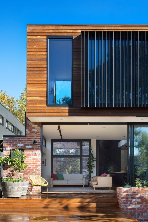 A sunny two-storey contemporary residential facade with warm horizontal timber cladding, exposed red brickwork, dark-framed glazing, and a prominent vertical metal screen over the upper-level window. The open ground-floor living area connects to a timber deck with built-in brick planters and outdoor seating. The image emphasizes the facade, glazing, materials, and indoor-outdoor connection.