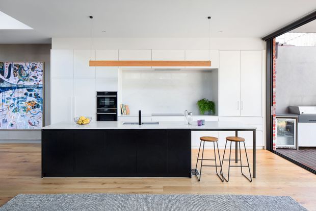 Bright contemporary kitchen with a large black island, white full-height cabinetry, integrated appliances, a pale stone benchtop, timber pendant light, and two bar stools. Timber flooring, colourful artwork, and a fully glazed opening to an outdoor barbecue area are visible.