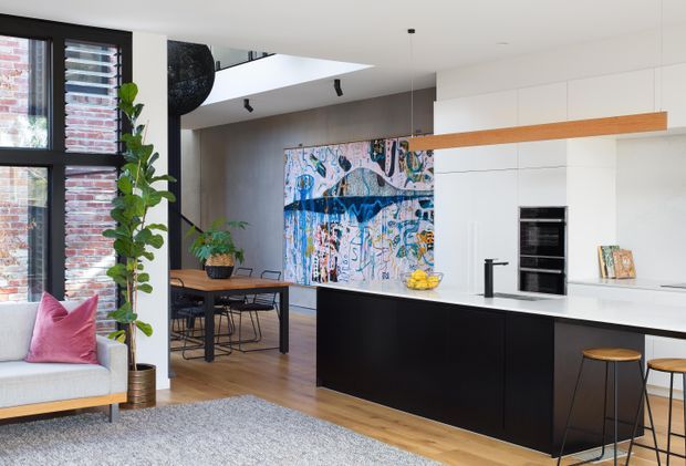 Bright open-plan kitchen and dining area with a black island, white full-height cabinetry, integrated appliances, timber flooring, and a timber feature beam. A large colourful artwork defines the dining wall, while a light lounge chair, indoor plants, and black-framed glazing add contrast. The room has a contemporary, refined feel with a visible high ceiling and partial double-height void.