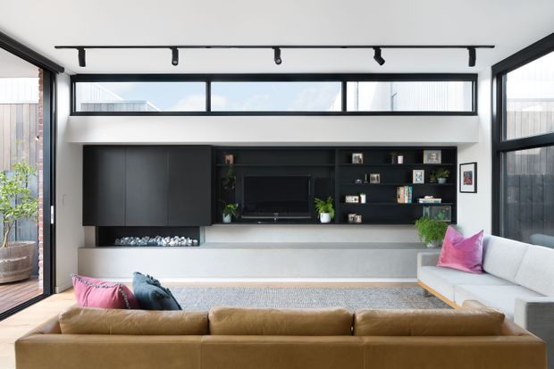 Bright contemporary living room with a large tan leather sectional sofa facing a dark built-in media wall. The joinery incorporates a television, open shelving, decorative objects, plants, and a long horizontal fireplace with a pale stone or concrete ledge. Black-framed floor-to-ceiling glazing and high clerestory windows bring in abundant daylight, with track lighting across the ceiling and a glimpse of an outdoor courtyard at left.
