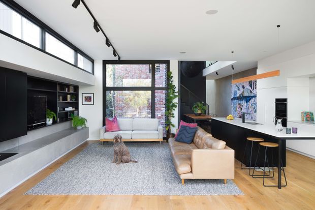 Open-plan contemporary living room with a visible kitchen and dining counter. The room has light timber flooring, a large grey rug, a tan leather sofa, a pale sofa beneath expansive black-framed glazing, and extensive black built-in media and kitchen cabinetry. A high white ceiling includes track lighting, while a colourful artwork provides a small accent. The space feels bright, refined, and inviting.