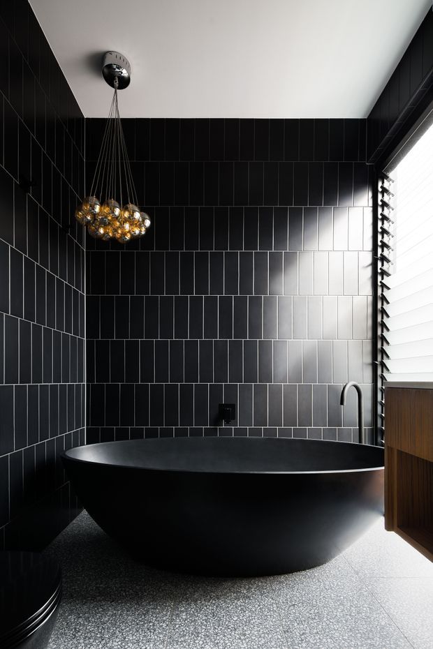 A dramatic contemporary bathroom featuring a large matte-black freestanding oval bathtub, black vertically stacked rectangular wall tiles with contrasting grout, a dark textured floor, and a sculptural pendant light with amber glass globes. A black wall-mounted tap and curved black bath filler sit beside the tub, while a louvred window brings in strong daylight and a dark timber vanity is partially visible at right.