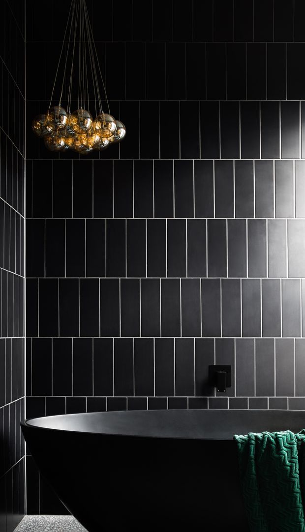 Moody, dramatic bathroom interior featuring a freestanding matte-black bathtub against full-height black rectangular wall tiles with contrasting light grout. A sculptural cluster pendant with amber glass globes hangs above the bath, while a deep green textured towel provides the main accent. The space has dark, refined finishes and strong directional lighting.