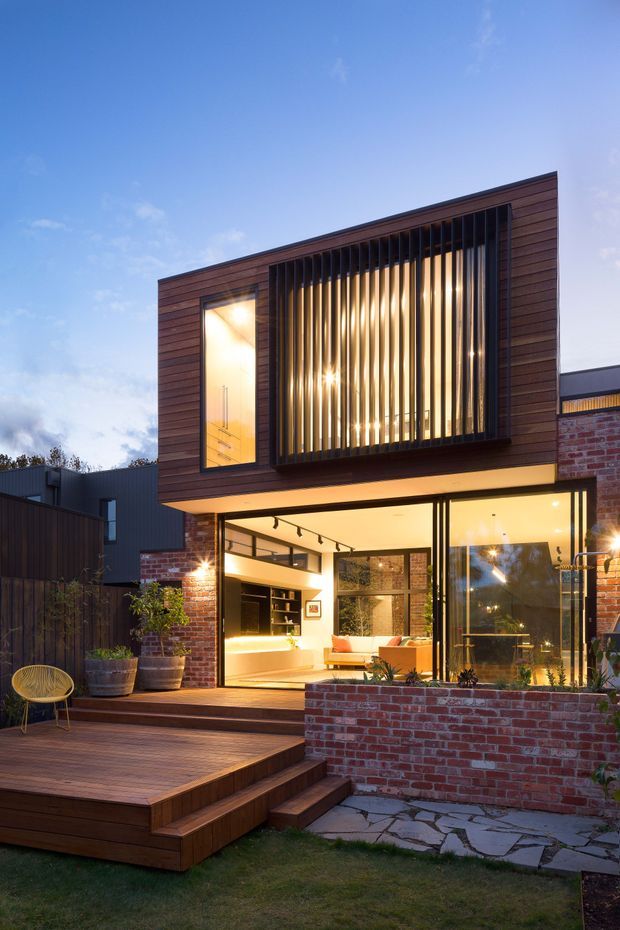 Twilight exterior view of a contemporary two-storey urban residence. The façade combines warm horizontal timber cladding, exposed red brick and extensive floor-to-ceiling glazing. A prominent upper-level window is screened with vertical dark metal or timber fins, while the illuminated ground floor opens onto a broad timber deck with steps, brick planters and a stone-paved garden path.