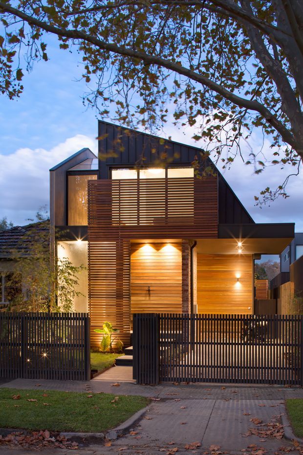 Photograph of a contemporary multi-level urban residence at dusk, viewed from the street behind a dark vertical-slat fence and gate. The facade combines warm timber horizontal screening and cladding with dark standing-seam metal-like wall panels, large glazed openings, and a sharply pitched asymmetrical roof form. Warm integrated lighting illuminates the entry, timber walls, and covered carport or porch area. Mature tree branches frame the upper foreground, with a paved driveway and small landscaped front garden visible.