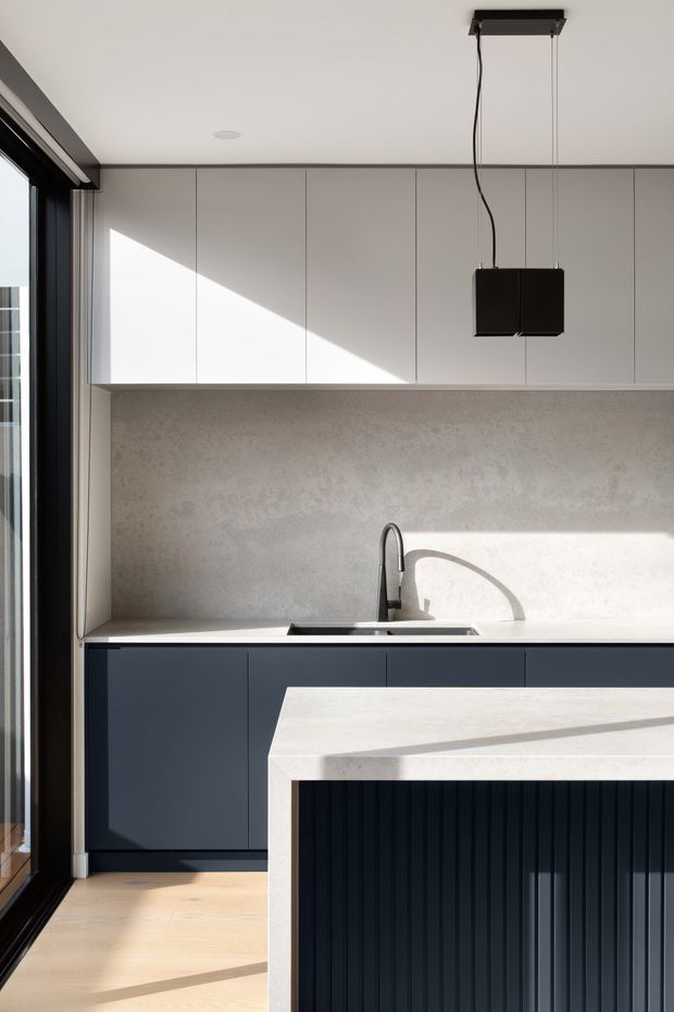 A clean, contemporary kitchen photographed in strong natural light. The compact galley-style kitchen has flat-panel matte dark navy lower cabinetry, pale grey-white overhead cabinets, a continuous light stone-look benchtop and matching splashback, a dark integrated sink and slim black tap, and a pendant light above the island. The island features a thick pale benchtop with a dark vertically fluted front. Full-height dark-framed glazing is visible to the left, and light timber flooring runs through the space.