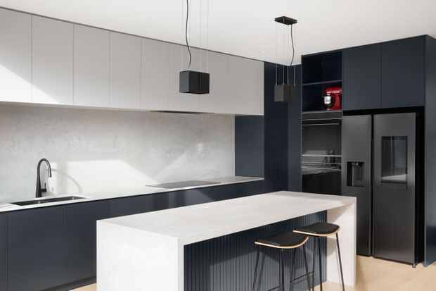 Contemporary kitchen with a large white waterfall-edge island, dark navy-black cabinetry, integrated appliances, pale upper cabinetry, a light stone-look splashback, and two suspended geometric pendant lights. The island includes a vertically ribbed dark feature panel and seating for two. Light timber flooring adds warmth to the otherwise monochromatic palette.