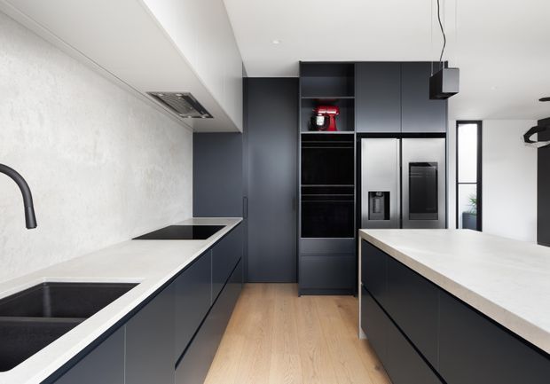 A contemporary galley-style kitchen with a central island. The space features dark matte flat-panel cabinetry, pale stone-look benchtops, integrated stainless-steel appliances, a black undermount sink and tap, light timber flooring, and a white textured wall finish. Minimal black pendant lighting and open shelving add contrast.