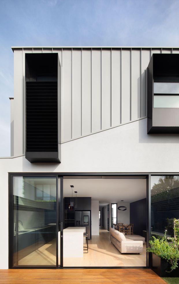 A contemporary multi-level residential facade viewed from a timber deck. The upper facade combines pale vertical metal panel cladding with a smooth white wall, deep black-framed projecting window openings, and a dark recessed vertical louvre feature. Large sliding glass doors open into a bright open-plan interior containing a kitchen island, living area, dining table, and timber flooring. A small planted area borders the deck.