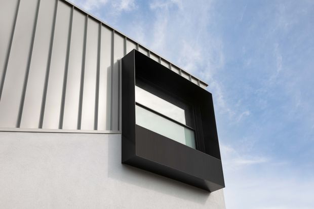 Close-up exterior view of a contemporary building facade, featuring a crisp white rendered wall, vertically ribbed light metal cladding, and a projecting black-framed upper-level window or architectural box. The composition emphasizes strong geometric lines, contrasting materials, and modern detailing against a blue sky.