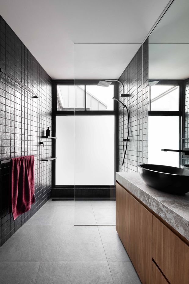 A contemporary bathroom with a walk-in glass shower, full-height black grid-pattern wall tiles, black-framed windows, a wall-mounted black shower system, and a floating timber vanity with a grey stone benchtop and matte black vessel basin. Large grey floor tiles and a burgundy towel add contrast, while the mirrored wall visually extends the narrow space.