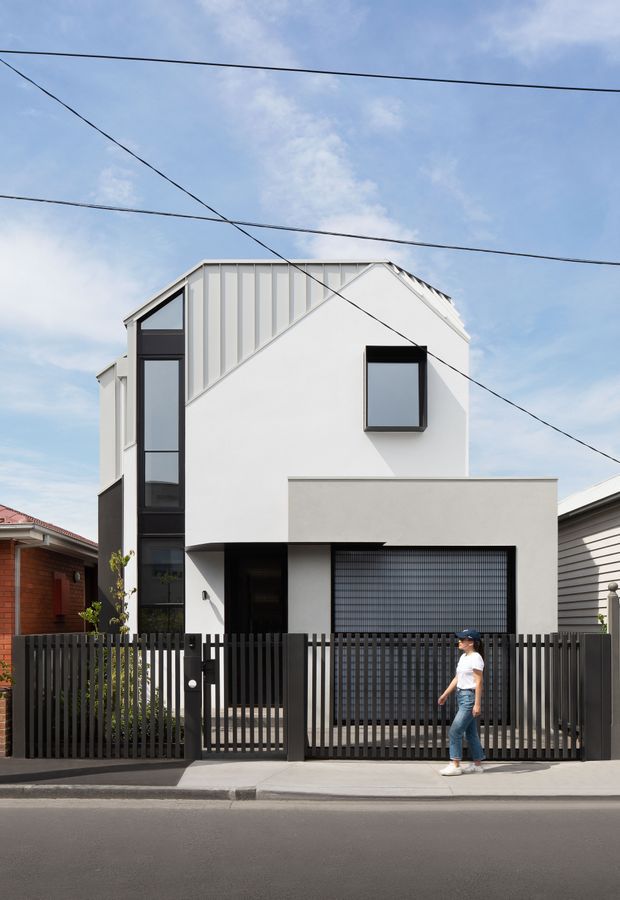 Street-facing photograph of a narrow multi-storey urban residence with a crisp white rendered facade, dark vertical window framing, a pitched gable roof, and a projecting ground-floor garage volume. A black vertical-slat fence and sliding gate enclose the frontage, with neighbouring homes and overhead utility wires visible.