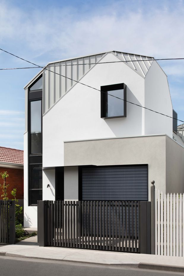 Street-facing photograph of a narrow contemporary multi-storey townhouse facade. The building has a steep gabled roof with folded standing-seam metal cladding, white rendered walls, a projecting dark-framed upper window, tall vertical glazing, and a ground-level garage with a dark gridded door. A black vertical-slat security gate and fence define the frontage, beside a white picket fence. Overhead utility wires cross the view.