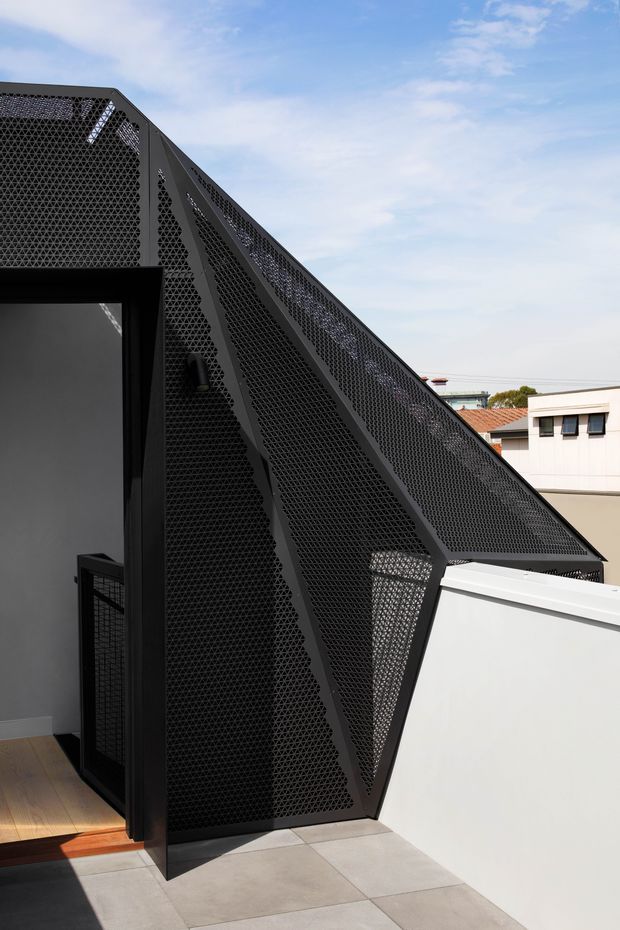 Exterior architectural photograph of a contemporary rooftop or upper-level structure featuring a sharply angled, triangular black metal screen facade with fine perforated geometric mesh. The screen forms a dramatic pitched or folded roof-like profile beside a dark-framed opening. A white parapet wall, pale tiled terrace, neighbouring buildings, and blue sky are visible.
