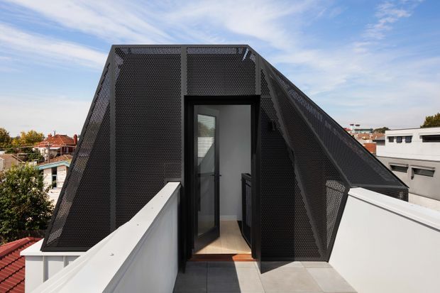 Exterior rooftop view of a distinctive dark perforated-metal enclosure with steeply sloping sides and a central glazed door, accessed from a light-coloured terrace or balcony. The structure forms a strong geometric, angular silhouette against a suburban urban backdrop and blue sky.