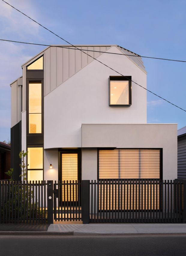 Street-facing view of a narrow contemporary multi-storey residence at dusk. The facade combines smooth white rendered surfaces with vertical metal-clad upper sections, tall black-framed windows, illuminated interiors, and a projecting ground-floor volume. A dark vertical-bar security fence and gate enclose the frontage, with neighbouring homes and overhead utility wires visible.