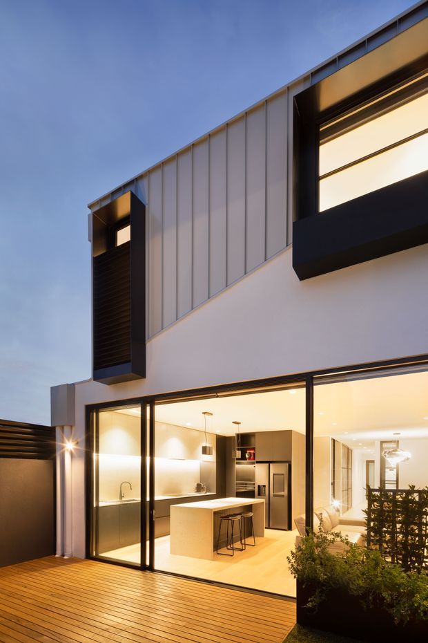 Twilight exterior view of a contemporary multi-storey residence with a white rendered lower facade, vertically folded metal upper cladding, dark framed windows, and projecting black window surrounds. Full-height glazing opens onto a timber deck and reveals a warmly lit open-plan kitchen with a central island. A dark privacy screen and planted garden bed frame the foreground.