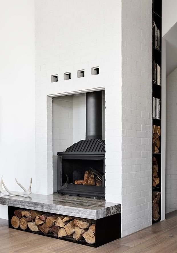 Interior view of a contemporary living area centred on a black wood-burning fireplace set within a white-painted brick chimney breast. The fireplace has a substantial grey stone or concrete mantel and hearth, with an open firewood storage niche below and a tall integrated firewood shelving bay to the right. Small square ventilation openings punctuate the chimney breast, while dark timber flooring and minimal styling create a clean, modern composition.