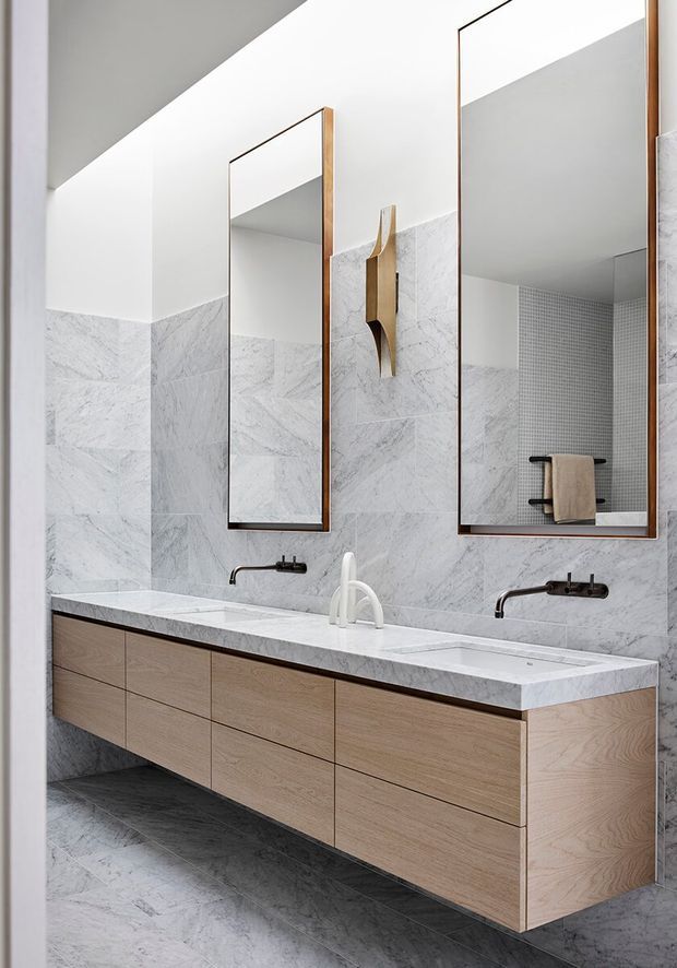 A bright contemporary bathroom featuring a long floating double vanity in light timber cabinetry, a white marble-look stone benchtop and integrated basins, wall-mounted black tapware, large frameless mirrors with timber edging, and pale marble-look wall and floor tiles. A small tiled shower area with a towel rail is visible in the reflection.