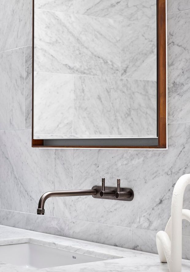 Close-up of a refined bathroom vanity and bathtub area clad in white veined marble. A rectangular timber-framed mirror hangs above a wall-mounted brushed dark-metal tap with separate controls. The marble extends across the wall and countertop, with a white sculptural object partially visible at right.