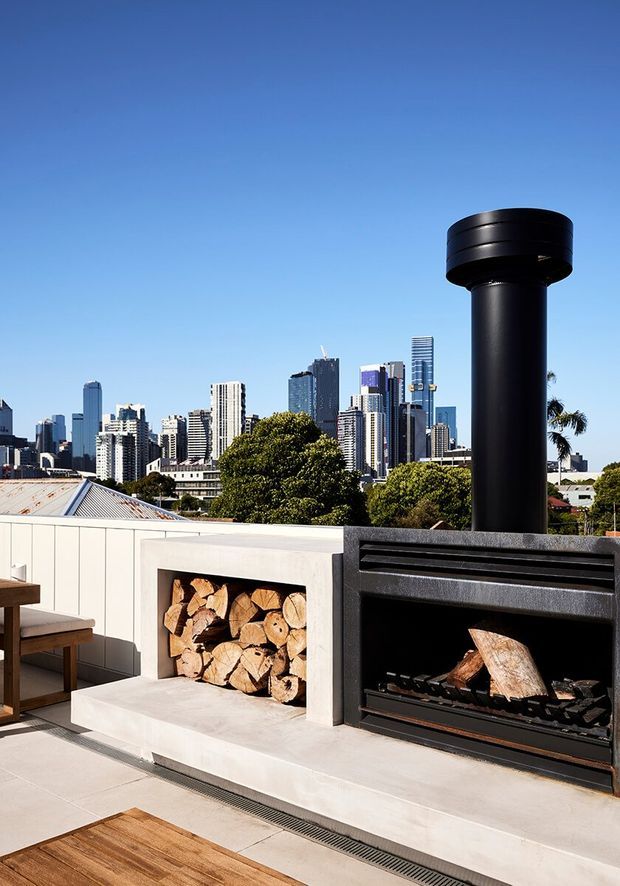 A bright rooftop or terrace outdoor living area overlooking a dense modern city skyline. The foreground features a substantial black cylindrical flue connected to a built-in black fireplace, adjacent to an open white-rendered wood-storage niche filled with split firewood. Pale tiled or concrete paving, timber furniture, and planted trees frame the urban view under a clear blue sky.