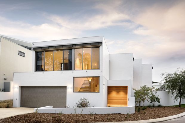 A contemporary multi-level residential facade with crisp white masonry walls, a flat roofline, expansive upper-level glazing, a large integrated garage, and a warm timber-lined recessed entry or garage opening. The home sits on a landscaped suburban street frontage with a minimalist hardscape and sparse planting.