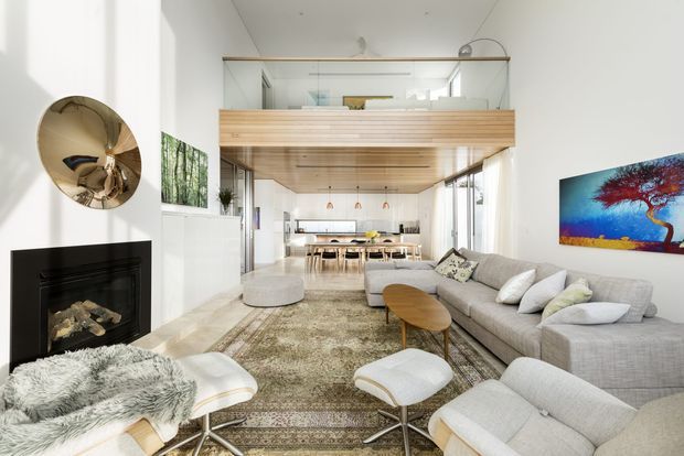 Bright open-plan living and dining space with a double-height white interior, large grey sectional seating, patterned area rug, fireplace, and a timber-lined ceiling feature extending over the dining area. Full-height glazing brings in abundant natural light, while a colourful artwork and sculptural wall mirror add accents.
